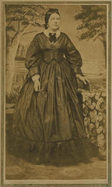 Carte-de-visite full-length portrait in front of a painted backdrop of Mrs. Belle Knickerbocker of Shullsburg, Wisconsin. She is holding a book in her left hand that is resting on a cloth covered table.