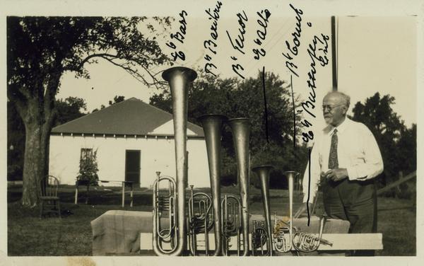 A man posing outdoors with musical horns, with notations: "E flat bass, B flat Baritone, B flat Tenor, E flat Alto, B flat Cornet, E flat 'Butterfly' Cornet".