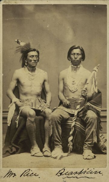 Full-length seated portrait of Henry Rice and Bearskin (Hoon-jah-gah), both of the Ho-Chunk (Winnebago) tribe.  Bearskin was a member of the Bear Clan and a leader in the medicine lodge.  Bearskin's son, John, lived in Nebraska.