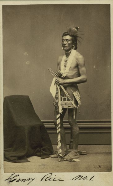 Full-length portrait of Songjonmanneekaw (Henry Rice) of the Ho-Chunk (Winnebago) tribe posed standing next to a chair covered with a cloth.  Songjonmanneekaw was the son of Chief Little Hill and was a renowned medicine man.  He died around 1911 in Wisconsin.