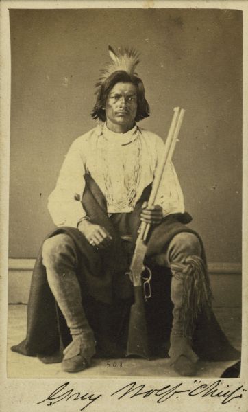 Portrait of Grey Wolf, brother of Little Priest, sitting and holding a rifle.