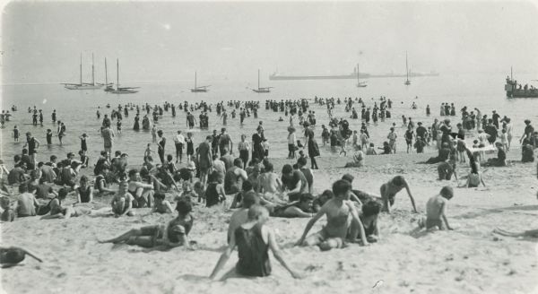 Men, women and children soaking up the sun on the beach of a lake (probably Lake Michigan). The children are digging in the sand in the foreground, and there are ships in the distance.