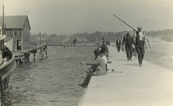 Pier lined with people fishing on a lake (probably Lake Michigan). Men are returning toward shore, and other men are sitting and fishing, lined up along the length of the pier. In the left foreground is a boat, and across the water on the left is a building suspended over the lake by posts.