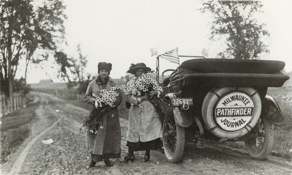Rear view of Robert Taylor's car, which he used for his work as the <i>Milwaukee Journal</i>'s first photographer. A sign displayed across the spare tire reads "<i>Milwaukee Journal</i> Pathfinder," an allusion to the Journal's highway information department.  Two women, one of whom is Taylor's wife Alma, hold armloads of flowers.  The car is stopped along a rural road.