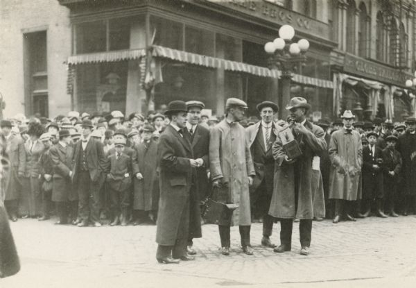 Five men stand on a city street surrounded by onlookers, perhaps a parade or some other public event. Two of the men in the street are carrying photography equipment.