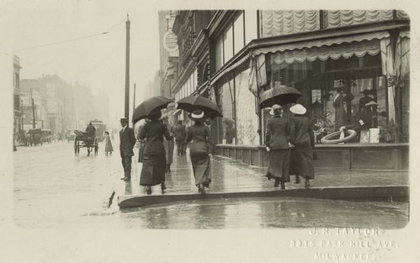 A street scape with people holding umbrellas walking on a sidewalk in the rain. The street shows a business section, and there is a horse-drawn vehicle in the hazy distance.