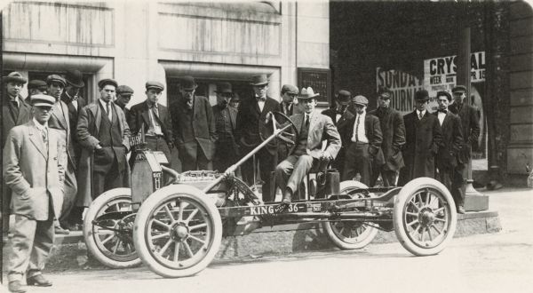 View towards a man sitting in an open car parked on the street. The driver in the vehicle is surrounded by interested onlookers. The left side of the car has a sign that reads: "King - Silent - 36," "A Thousand Less Parts" and "Easy For Riding."