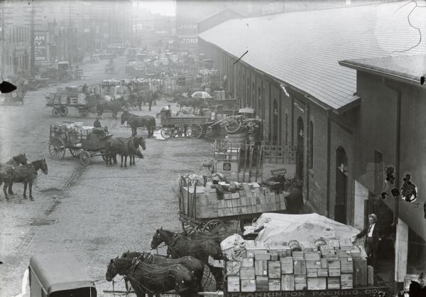 Elevated view of the industrial warehouse district. There are horse-drawn wagons lined up along loading docks, with men working in the area.