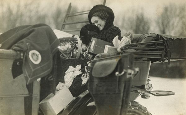 A woman dressed for winter weather and seated in the back of a touring car, looks coyly at the photographer. She is surrounded by holiday gifts and packages.