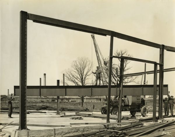 A construction site with a truck manufactured by Winther Motor and Truck Company. A forty foot boom is mounted on the truck. Workers are positioning a steel beam.
