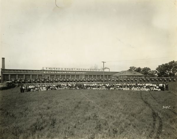 Exterior of Winther Motor and Truck Company factory with a row of trucks in front, and workers posing in front of the trucks.