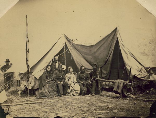 A group of people at the headquarters tent of the 2nd Wisconsin Infantry. Seated (left to right) are officers Lucius Fairchild, Colonel Edwin O'Connor, and Major Thomas Allen. With them are Mrs. Hathaway, Captain Hathaway (standing) and Mary Howe. Behind the tent is a man identified in Fairchild's own handwriting as Ralph, who wears one of the black hats for which the 2nd Wisconsin and other regiments in the Iron Brigade were famous.