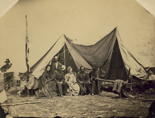 A group of people at the headquarters tent of the 2nd Wisconsin Infantry.  Seated (left to right) are officers Lucius Fairchild, Colonel Edwin O'Connor, and Major Thomas Allen. With them are Mrs. Hathaway, Captain Hathaway (standing) and Mary Howe. Behind the tent is a man identified in Fairchild's own handwriting as Ralph, who wears one of the black hats for which the 2nd Wisconsin and other regiments in the Iron Brigade were famous.