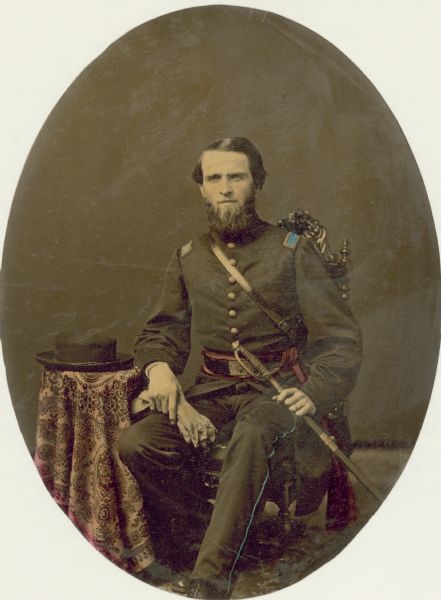 Full-length seated studio portrait of Second Lieutenant Charles J. Robinson of the First Regiment of the Wisconsin Volunteer Infantry, G Company, commanded by Colonel John C. Starkweather. He is holding a sword on his left thigh, and a pair of gloves in his right hand. A hat is on the table next to his chair.