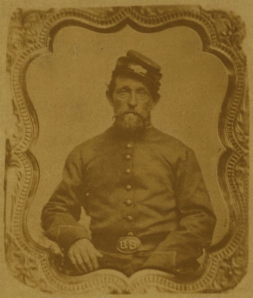 Waist-up seated portrait of an unnamed Union soldier. Cased frame around portrait. He is wearing a hat, and a US belt buckle.