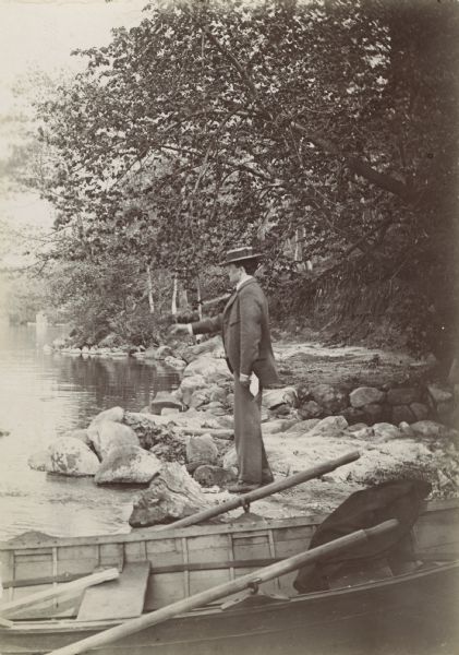 A man stands next to a rowboat on the rocky lake shore.