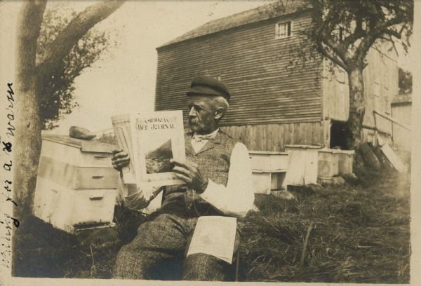 A bee farmer reads <i>America's Bee Journal</i> while sitting next to a number of hives.