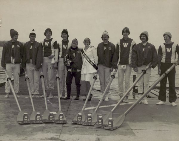 Wisconsin Honey Queen and nine members of the University of Wisconsin Madison's Crew Team next to Lake Mendota holding their oars with jars of honey on them.