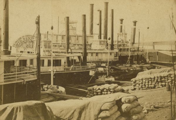 Steam-powered riverboats docked along the shoreline of a river.