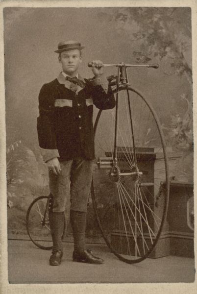 A carte-de-visite in front of a painted backdrop of a man standing next to a large bicycle.
