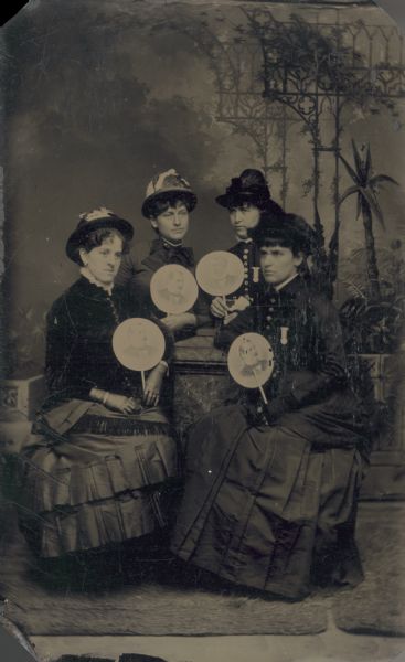 Studio group portrait tintype in front of a painted backdrop of four women sitting and holding hand fans with portraits of men printed on them. The women are wearing dresses and hats, and are posing with a prop stone wall.