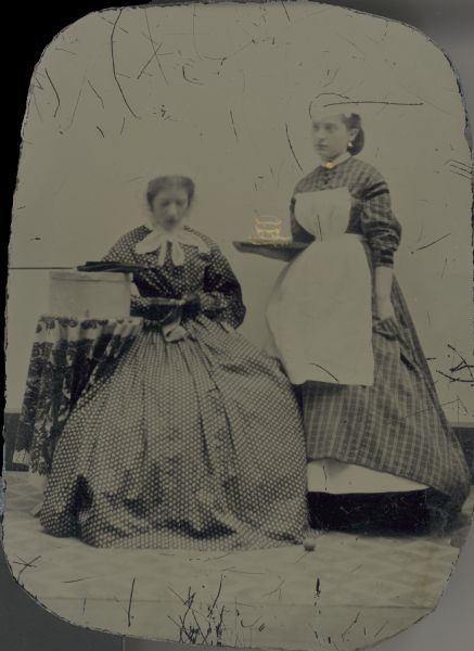 Tintype (ferrotype) of two women. One is seated and the other appears to be a maid, serving a drink.