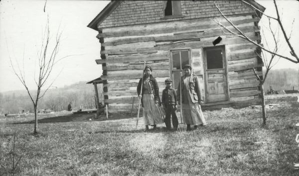 Three Indians of North America, two women and one boy, standing in front of a log cabin.