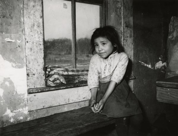 A young Menominee girl sitting next to a window.