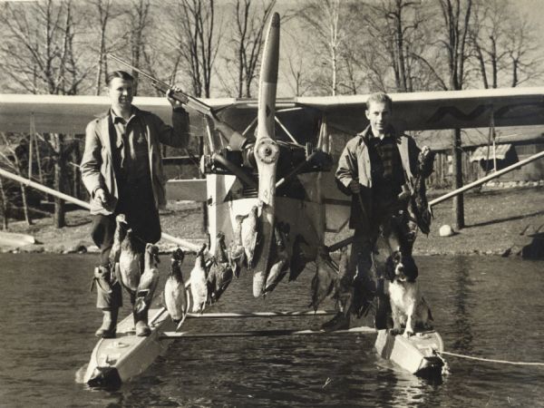 Two men standing on a pontoon airplane with their dog and a line of ducks they successfully hunted.