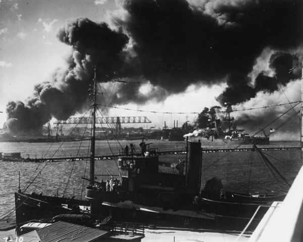 A tug or utility boat in the foreground and a battleship sit in Pearl Harbor with smoke rising in the background from the Japanese attack.