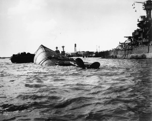Capsized USS <i>Oklahoma</i> at Pearl Harbor.  The <i>Oklahoma</i> was hit with a large number of Japanese aerial torpedoes which ripped an opening across most of the port-side hull plating of the ship, causing it to take on water and rotate into the water.  Sailors that were trapped in the ship were eventually cut free by other sailors and civilian dock workers at Pearl Harbor.
