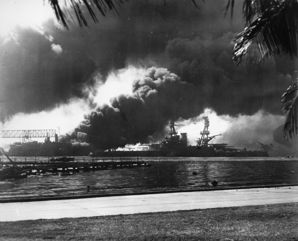 The USS <i>Nevada</i> in berth at Pearl Harbor with the USS <i>Shaw</i> burning in the background after its forward deck was hit with a Japanese bomb.