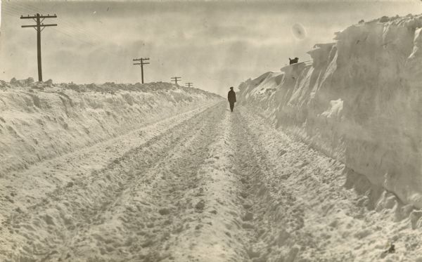 Winter scene with man walking down cleared train tracks, probably the Soo Line, next to man-sized drifts of snow, with a dog on the top of a drift. Between January 21-22, 1917, 9" of snow fell over western central Wisconsin with drifts up to 11.5 feet.