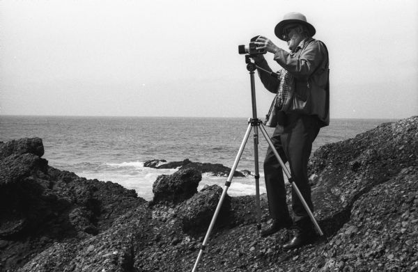 Photographer Ansel Adams mounting his Hasselblad camera onto the tripod on a rocky shore near or at Point Lobos.
