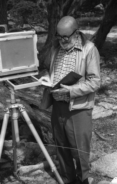 Photographer Ansel Adams standing near his 8 x 10 field camera and thumbing through his exposure record logbook in a wooded area near Point Lobos.