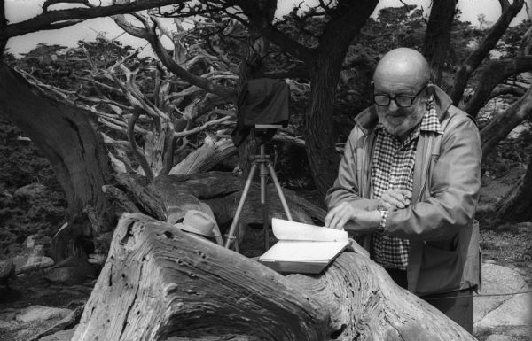 Ansel Adams checks the time as he records exposure information into his logbook during a photo shoot. His 8 x 10 field camera is in the background.
