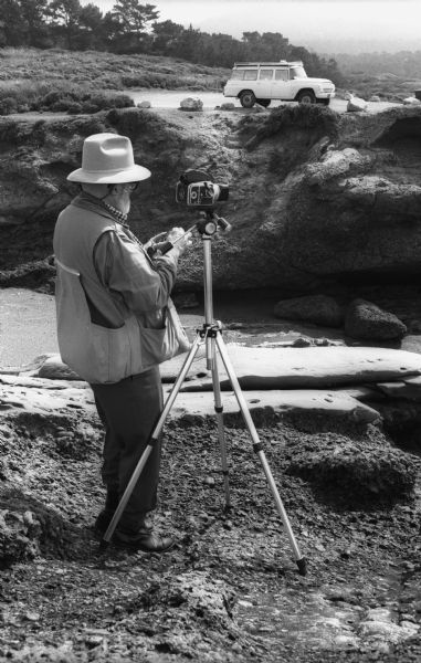 Ansel Adams takes a light reading with a hand-held light meter in front of his tripod mounted Hasselbald. His International Travelall is in the background.
