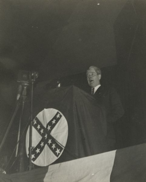 Governor Philip La Follette addressing the newly formed National Progressives of America at the University of Wisconsin-Madison Stock Pavilion. La Follette stands behind a podium draped with the Progressive Party symbol.