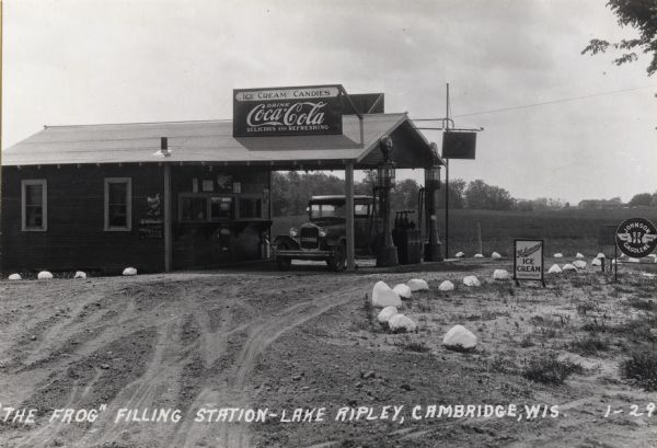 A Ford automobile parked at the "The Frog," a filling station between Cambridge and Lake Ripley that also offered motorists ice cream, soda pop, and candy.  This station was of the house and canopy type.