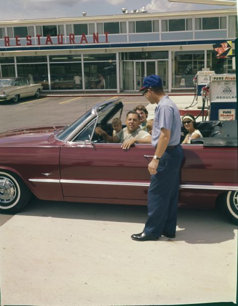 A family of travelers in a convertible asks directions of a gasoline attendant at a rest stop near Madison.