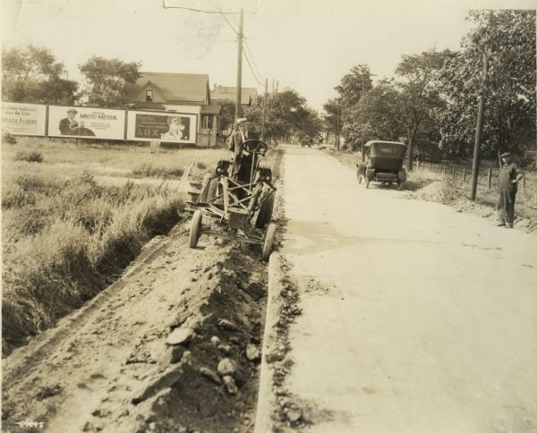 This photograph from the Wisconsin Highway Commission records is unidentified, but a few internal details helps to understand the content. The Wisconsin license plate on the automobile bears a 1922 date as well as a Wauwatosa identification. The Wauwatosa locale is confirmed by the billboards erected by the Cream City Bill Posting Company. The man driving the grading machine is obviously too well-dressed to be its regular operator. It is likely the regular operator is the man watching at the right, none too happy, while his supervisor, who had arrived in the car parked nearby, is testing a new piece of equipment.  At this time Milwaukee County was famous for its concrete roads.