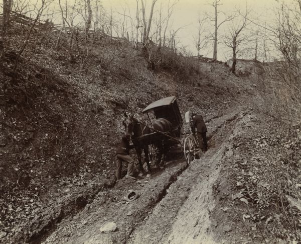 Two men attempt to move a buggy mired in the mud on a rutted, dirt road. One man is steadying the horse and his hat is laying in the middle of the road. The other man is trying to move a back wheel. This is one of a series of photographs collected by the League of American Wheelmen in 1898 from throughout the Midwest in order to publicize the need for better roads. (For another view of the same event see Image ID: 91862.)