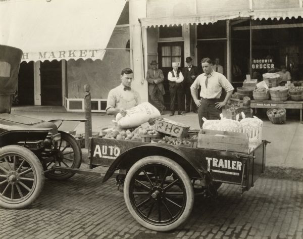 An auto-trailer, an early trailer manufactured by the Highway Trailer Company of Edgerton.  The company began by manufacturing trailers designed for farmers to haul goods to market using their cars.  When equipped with higher sides the trailer could be used for transporting cattle.  Behind the trailer is the Goodwin Grocery which identifies the location as brick-paved Vine Street in Beloit.