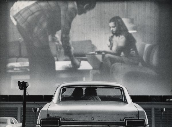 An evening at the Big Sky drive-in theatre. Rear view of two people sitting in an automobile watching a drive-in movie.