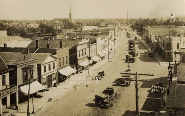 Elevated view of town. There is an unusual automobile parking pattern in the center of State Street. There is a sign for "Billiards" on a building in the right foreground. Other storefronts line the left side of the street. A large church is in the background.