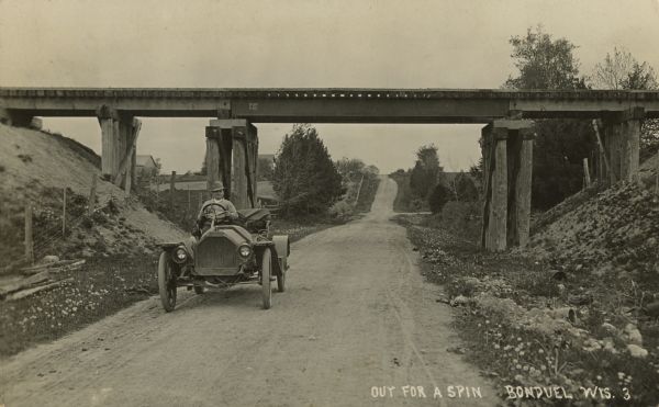 Photographic postcard showing a man driving an early open automobile with a railroad overpass behind him. Caption reads: "Out for a Spin, Bonduel, Wis."