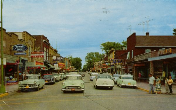 View down center of street in downtown Eagle River, with traffic, pedestrians, and automobiles parked along the curb. There are storefronts and signs on both sides of the street.