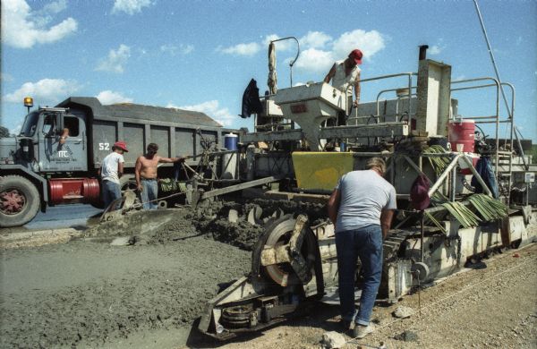Road construction workers at an unidentified location on I90 & I94 operate a construction machine that combines several steps of the concrete pour process. This was probably a demonstration that took place at an I90/94 "open house" sponsored by the Department of Transportation.