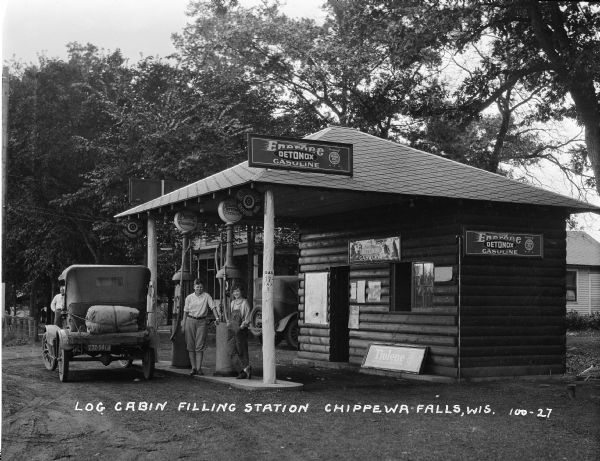 Rustic, log cabin gas station in Chippewa Falls with a traveler's vehicle being filled with gasoline. It is not clear if the young woman in overalls is a passenger or a local person or station employee. The latter seems likely, as the young man, while informally attired, is less casually dressed than the young woman.