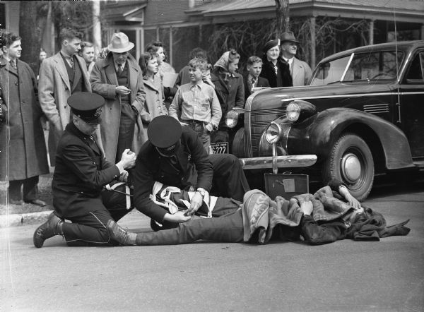 Stevens Point police officers apparently rendering first aid to a pedestrian injured by an automobile.  In fact, this photograph from the files of the State Office of Transportation Safety, represents a demonstration of accident response procedures at the annual State Safety Conference.